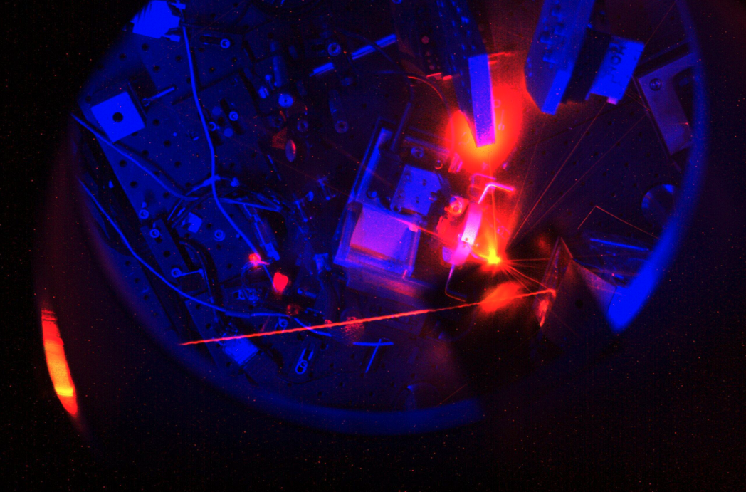 dark scene with blue hue and a bright red laser coming in from the left. The laser bounces off a mirror to the right of the image and appears to hit a target. Where it hits there is a bright red and yellow flare of light.